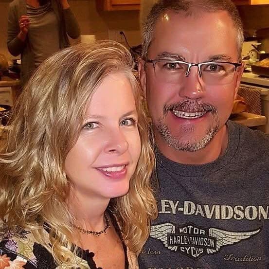 Smiling blonde woman and a man with glasses and a goatee posing closely together.