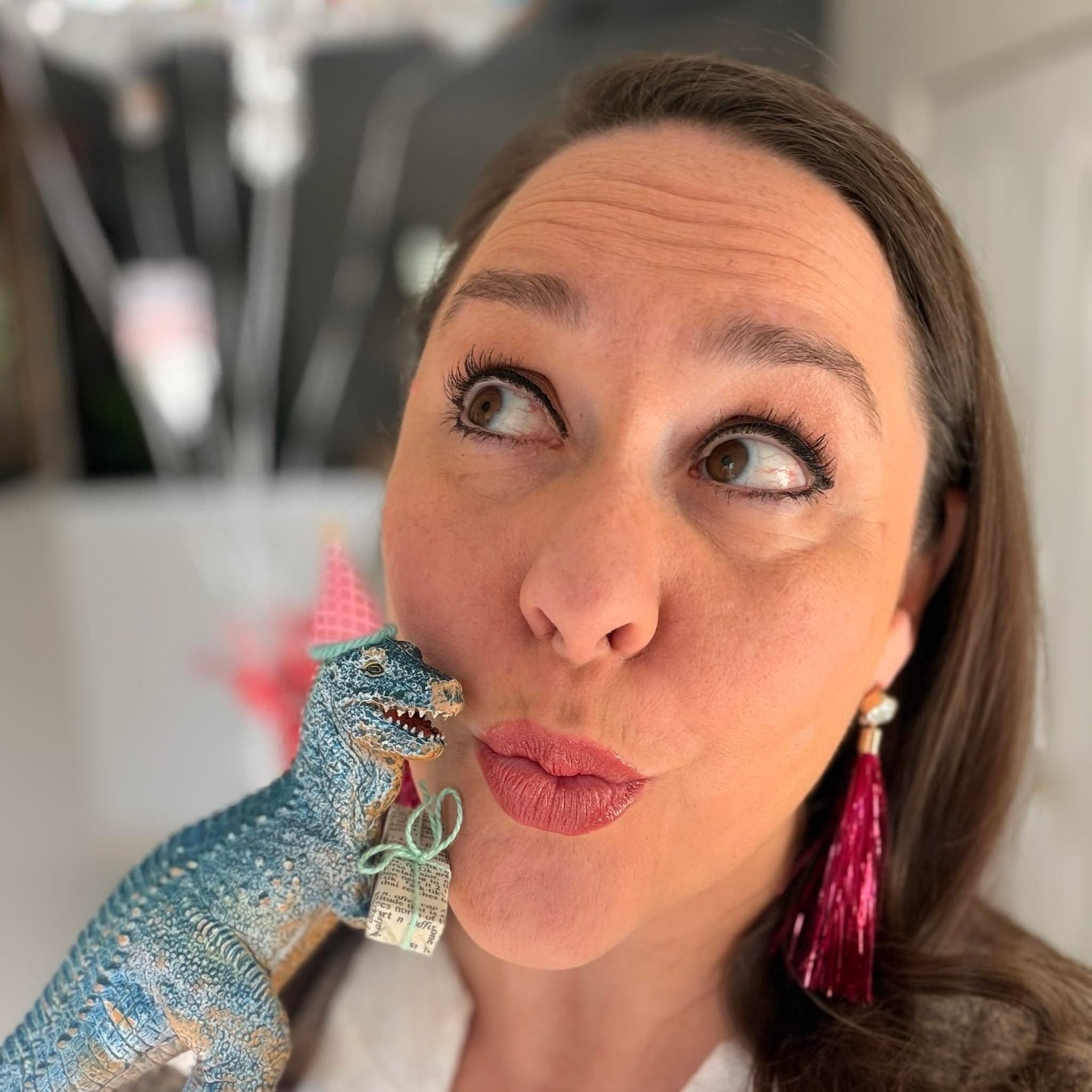 Playful woman looking up, holding a blue toy dinosaur wearing a tiny pink party hat.