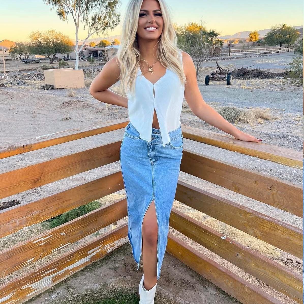 Smiling blonde woman wearing a white top and long denim skirt in a desert setting.